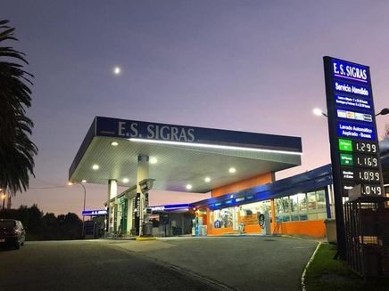 Gasolinera al anochecer, con marquesina iluminada, tienda de conveniencia y cartel de precios bajo un cielo que se oscurece