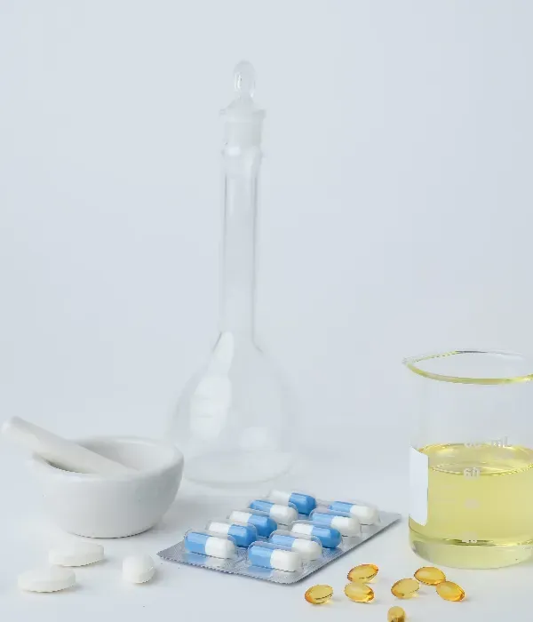 Sobre una mesa blanca, material médico: pastillas, cápsulas, mortero y maja, vaso de precipitados y frasco de vidrio.