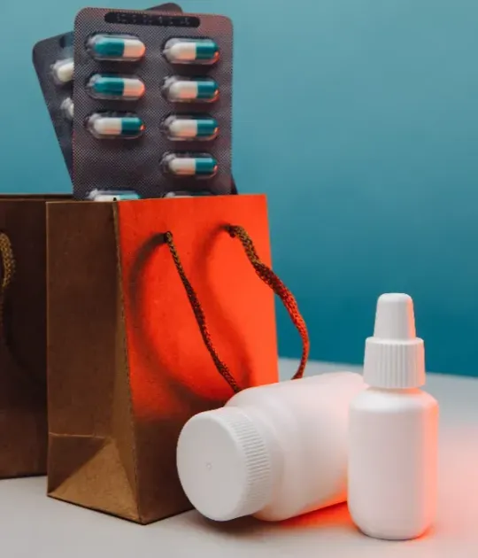 Frascos de medicamentos y blísteres en una bolsa de regalo marrón sobre un fondo verde azulado.