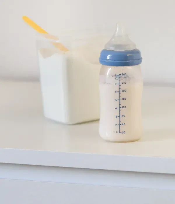 Un biberón lleno de leche de fórmula junto a un recipiente blanco con una cuchara medidora amarilla.