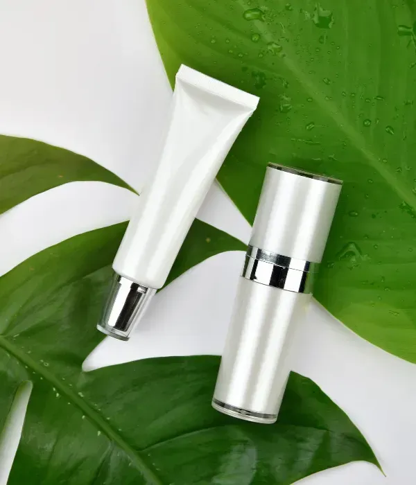 Tubo cosmético blanco y botella con tapa plateada sobre hojas verdes con gotas de agua.