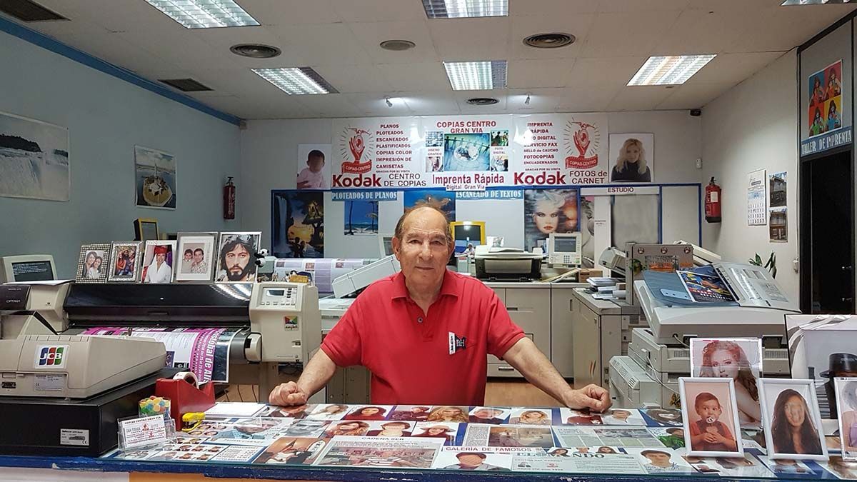 Un hombre con camisa roja se encuentra frente a un mostrador en una imprenta, rodeado de fotografías impresas y equipos.