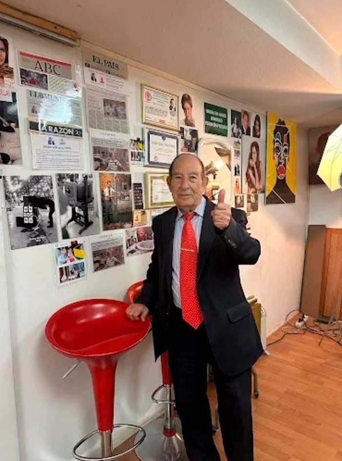 Un hombre de traje hace un gesto de aprobación con el pulgar cerca de un taburete rojo, con una pared decorada con fotos y premios.