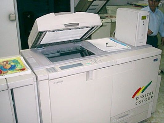 Fotocopiadora digital color Canon de gran tamaño, blanca, tapa abierta, documento sobre cristal, persona en el fondo.