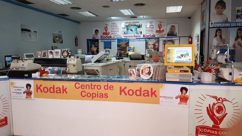 Un centro de copias Kodak con mostrador, equipo y carteles. Los colores amarillo, blanco y rojo son predominantes.