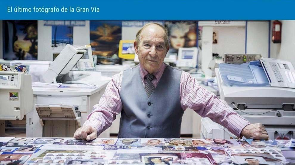 Un hombre mayor con chaleco en una imprenta, rodeado de materiales impresos, sonriendo.