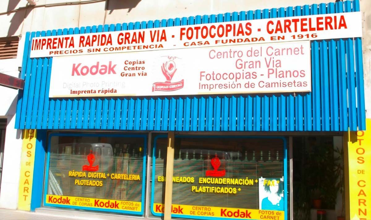 Una imprenta en la Gran Vía, España, con fachada azul y amarilla. Letreros de Kodak y diversos servicios anunciados.