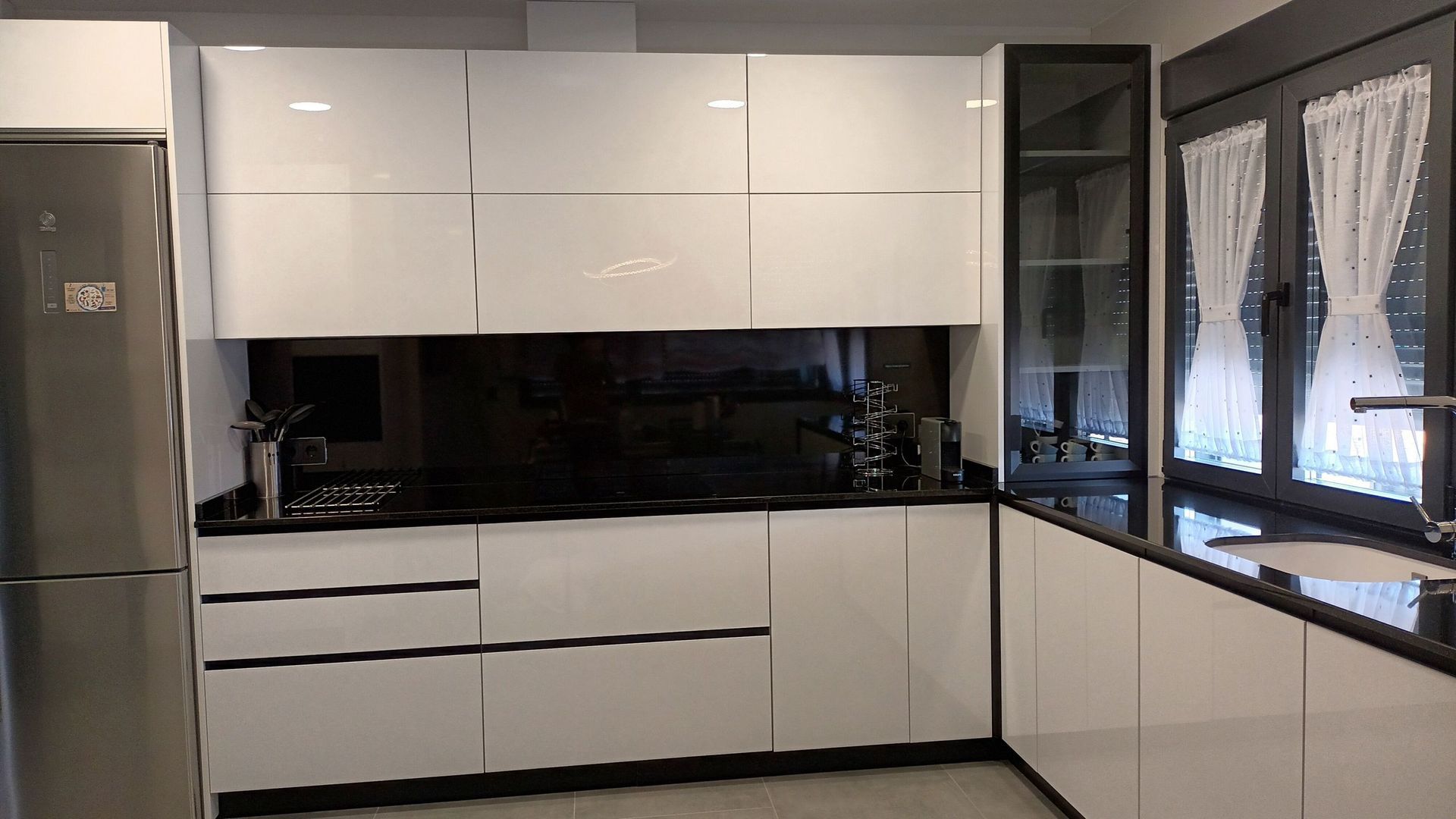 Una cocina moderna con armarios blancos brillantes, encimeras negras, un frigorífico de acero inoxidable y una ventana con cortinas.