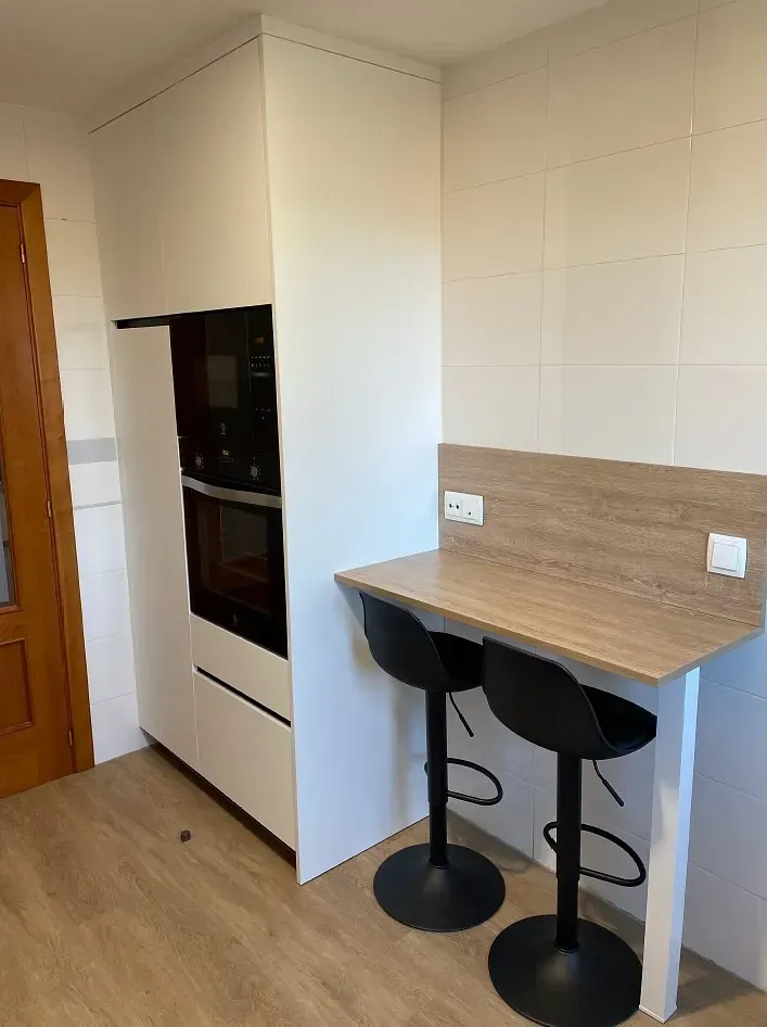 Un mueble de cocina alto de color blanco con horno y microondas integrados, situado junto a una barra de desayuno de madera con dos taburetes negros.