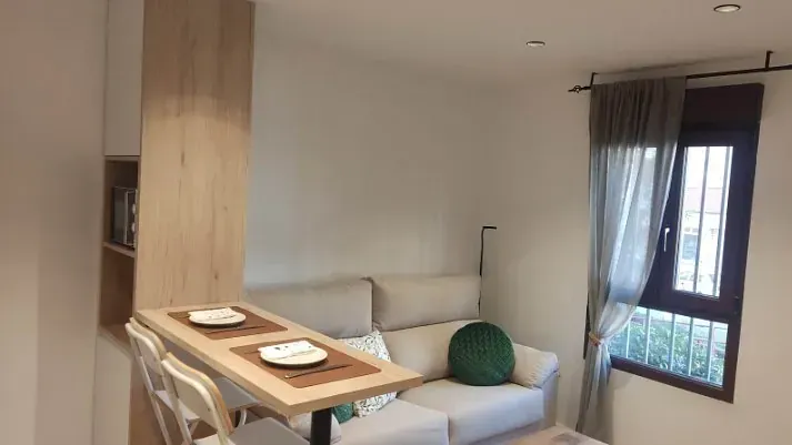 Un pequeño salón moderno con un sofá gris, una mesa de comedor de madera clara con dos sillas y una ventana con cortinas.