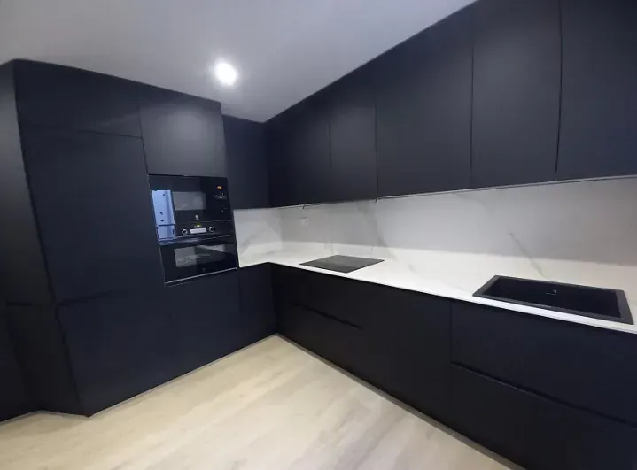Una moderna cocina en forma de L con armarios en negro mate, encimeras blancas y suelo de madera clara.