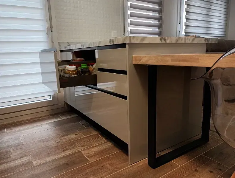 Una isla de cocina de color beige con cajones, uno de ellos parcialmente abierto, cuenta con una encimera de mármol y una barra de desayuno de madera clara.