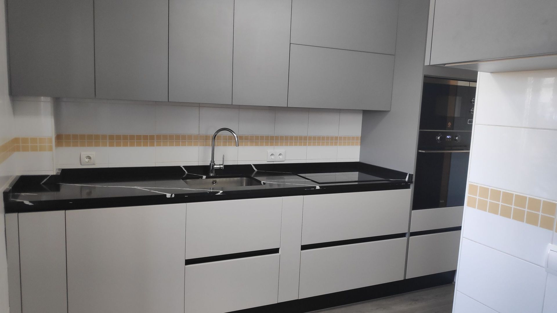 Una cocina moderna con armarios gris claro, encimera negra, fregadero de acero inoxidable y paredes alicatadas en blanco.