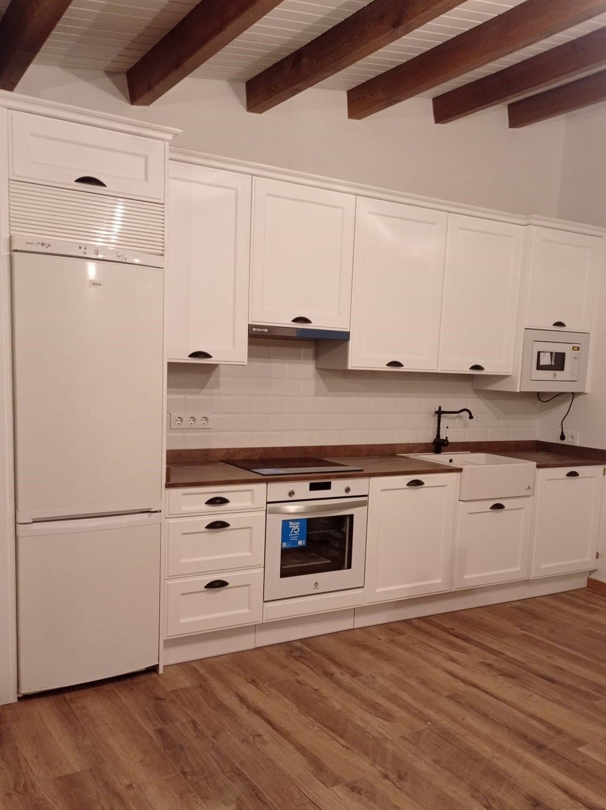 Una cocina moderna con armarios blancos, suelo con aspecto de madera, vigas de techo a la vista y electrodomésticos integrados de acero inoxidable.