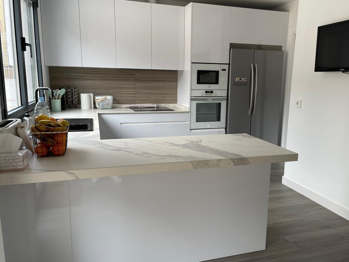 Cocina moderna con armarios blancos, encimeras de mármol, electrodomésticos de acero inoxidable y suelo de imitación madera gris.