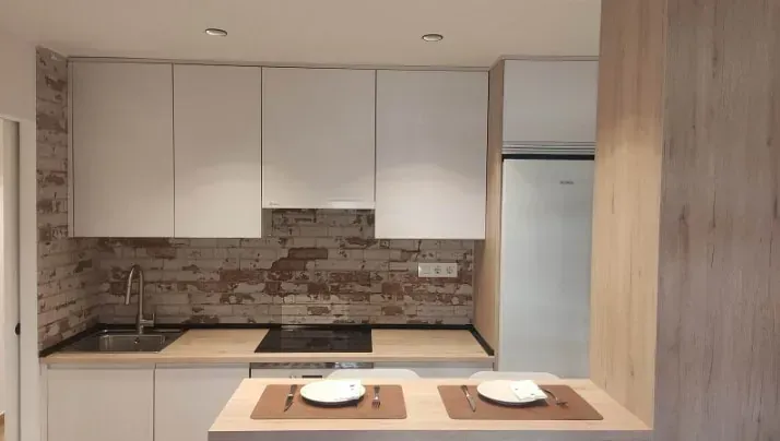 Cocina moderna con gabinetes blancos, encimeras en tono madera, salpicadero de ladrillo rústico y una barra de desayuno con dos servicios.