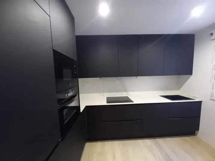 Una cocina moderna con armarios negros mate, encimeras blancas, electrodomésticos integrados y suelo de madera clara.