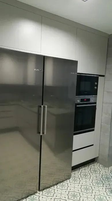Un gran refrigerador de acero inoxidable junto a un horno y un microondas empotrados en una cocina con piso de baldosas estampadas.