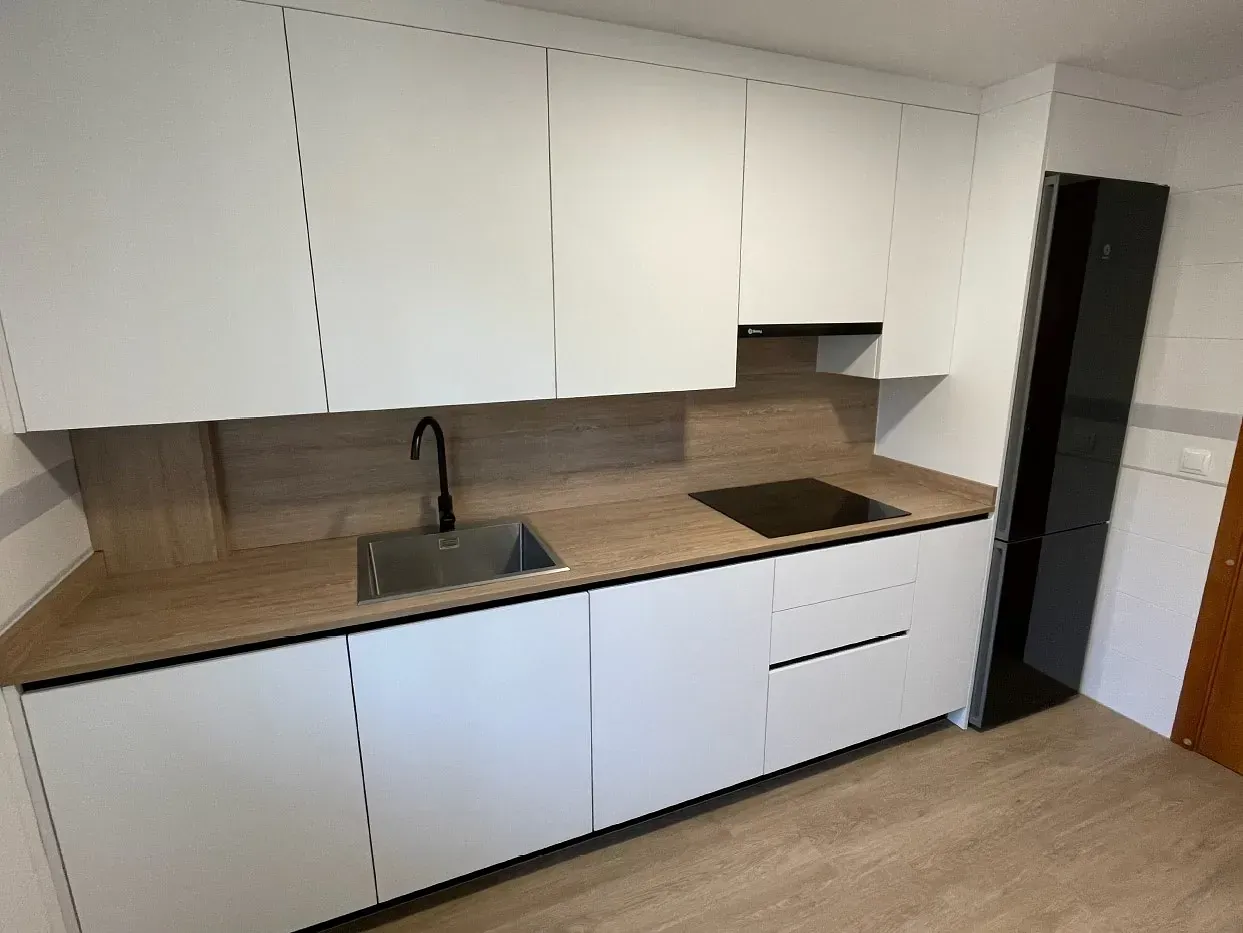 Una cocina minimalista con armarios blancos, encimera con diseño de madera, grifo negro y placa de cocción integrada negra.