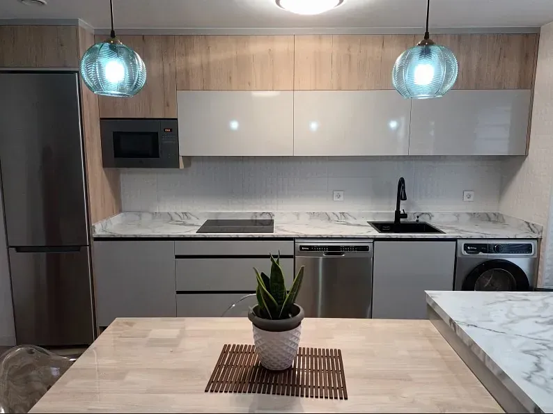 Una cocina moderna con detalles en madera clara, armarios blancos, electrodomésticos de acero inoxidable, encimeras de mármol y dos lámparas de cristal.