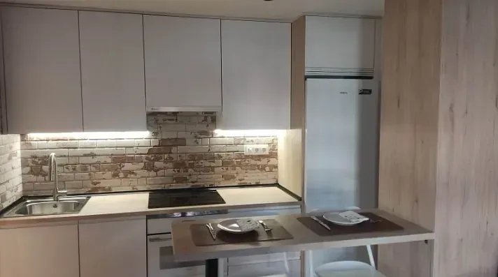 Una cocina moderna con armarios blancos, un salpicadero estilo ladrillo, una pequeña estufa, un frigorífico empotrado y una barra de desayuno.