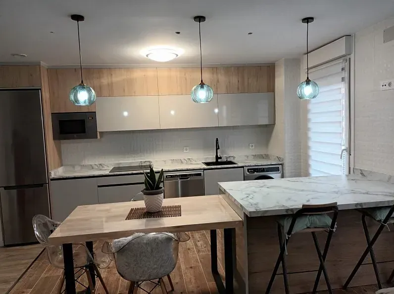 Una cocina moderna con armarios de madera clara, armarios superiores blancos, una isla de mármol y una mesa de comedor con tres lámparas colgantes.