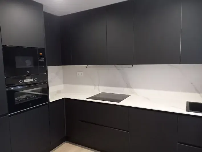Una cocina moderna con armarios negros mate, encimeras de mármol blanco, un protector contra salpicaduras blanco y electrodomésticos negros empotrados.