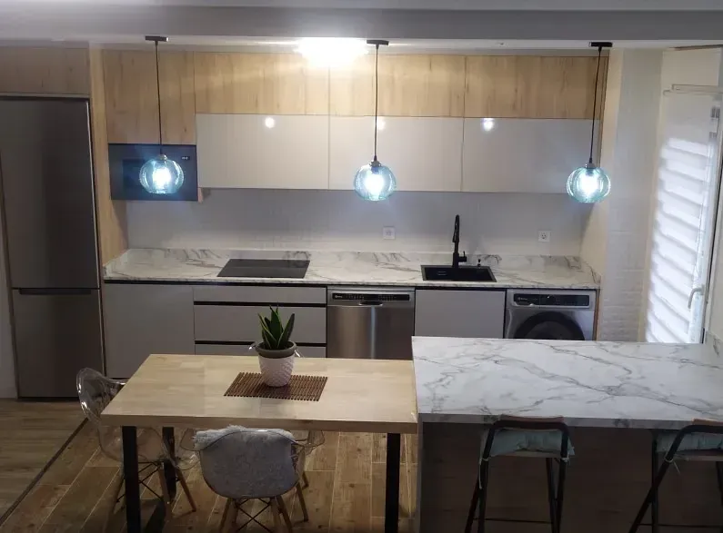 Cocina moderna con armarios de madera clara, armarios superiores blancos, encimeras con diseño de mármol y una mesa de comedor.