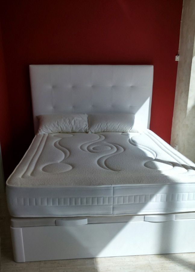 Una cama tapizada en blanco con cabecero capitoné y colchón texturizado contra una pared de color rojo intenso en una habitación soleada.