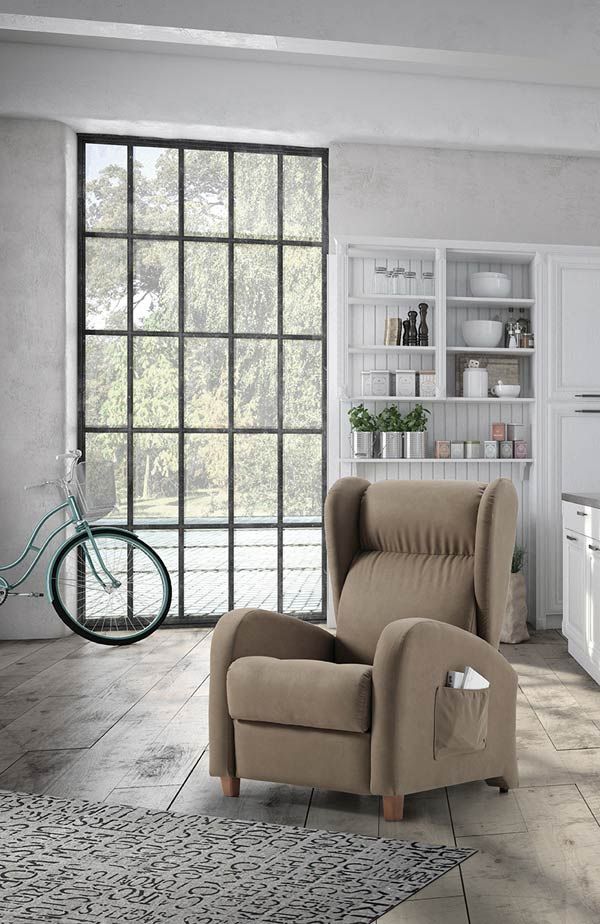 Un sillón reclinable de color beige con un bolsillo lateral descansa sobre un suelo de madera rústica en una habitación con una gran ventana enrejada y estanterías blancas.