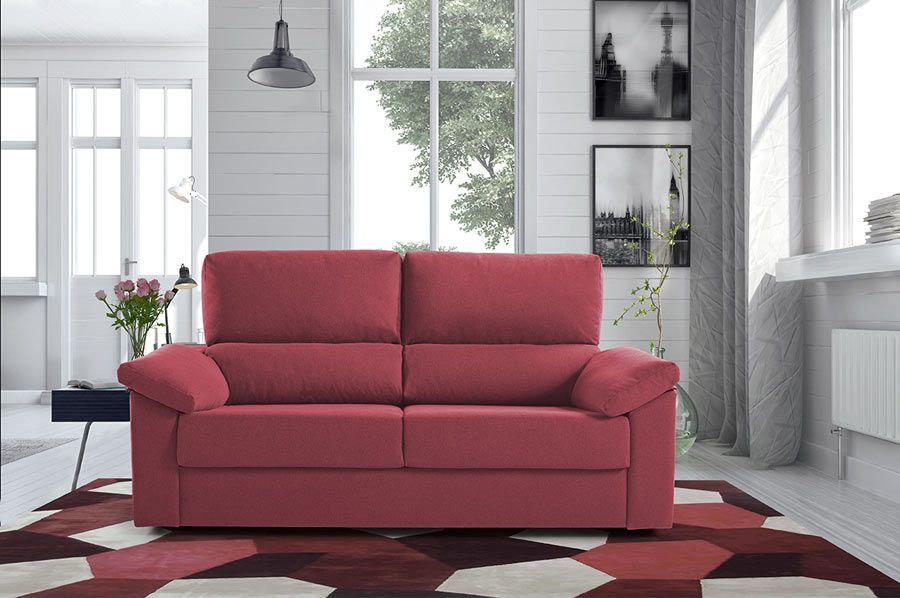 En una habitación luminosa, un sofá de dos plazas de color rosa se encuentra sobre una alfombra geométrica, centrado frente a una gran ventana.