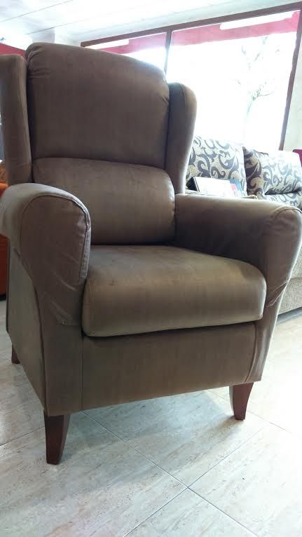 Un sillón orejero tapizado en color marrón con patas de madera oscura, situado en una sala de exposición muy iluminada.