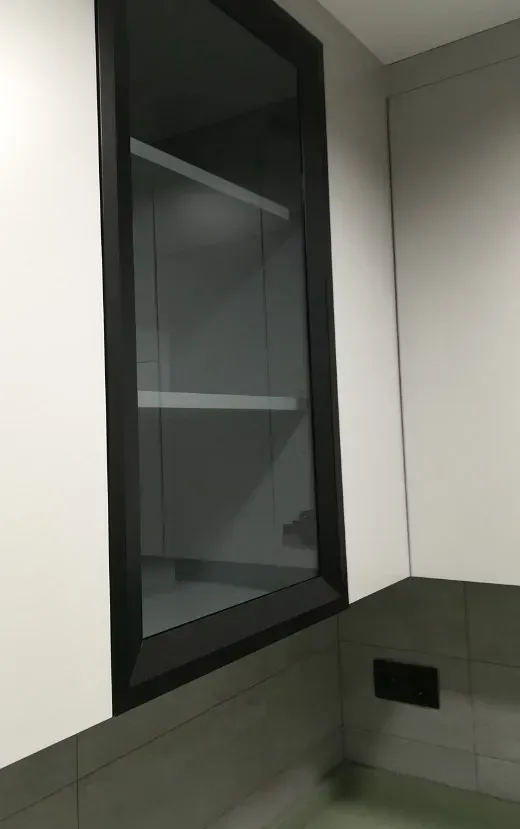 Un moderno armario vertical con marco negro y puerta de cristal, instalado sobre secciones de pared alicatadas en gris.