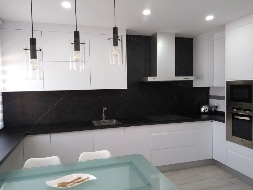 Una cocina moderna con armarios blancos, encimeras y salpicadero de piedra oscura, tres lámparas colgantes y una mesa de comedor.
