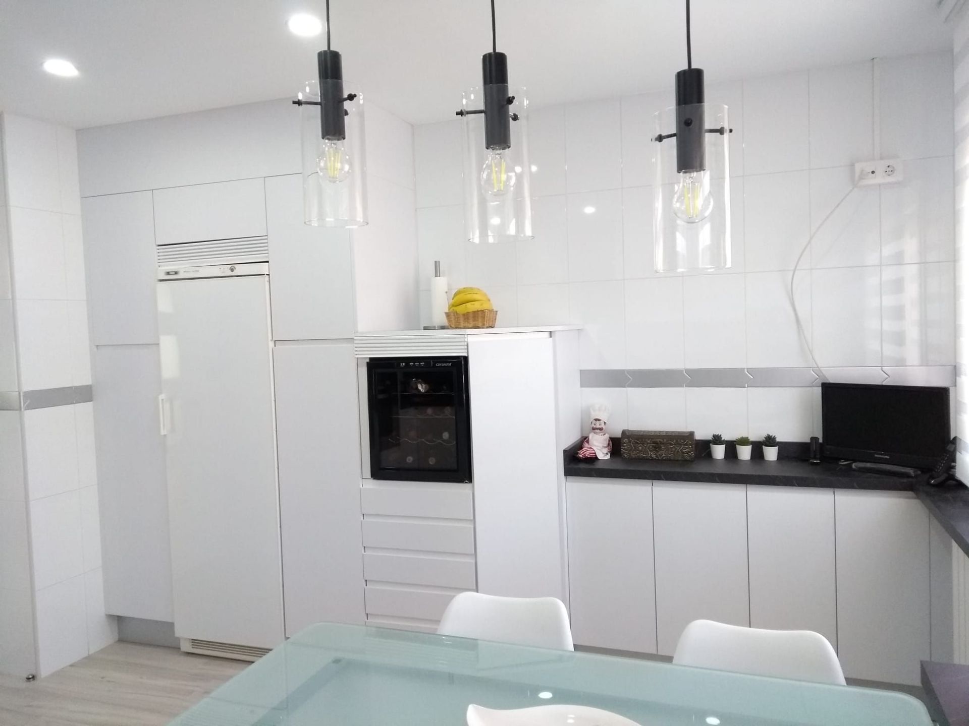Cocina moderna con armarios blancos, horno empotrado negro, lámparas colgantes y mesa con sillas blancas.