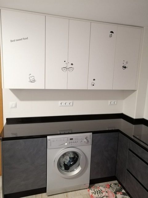Cocina moderna con armarios inferiores grises, armarios superiores blancos con pequeños adhesivos, lavadora y encimeras negras.