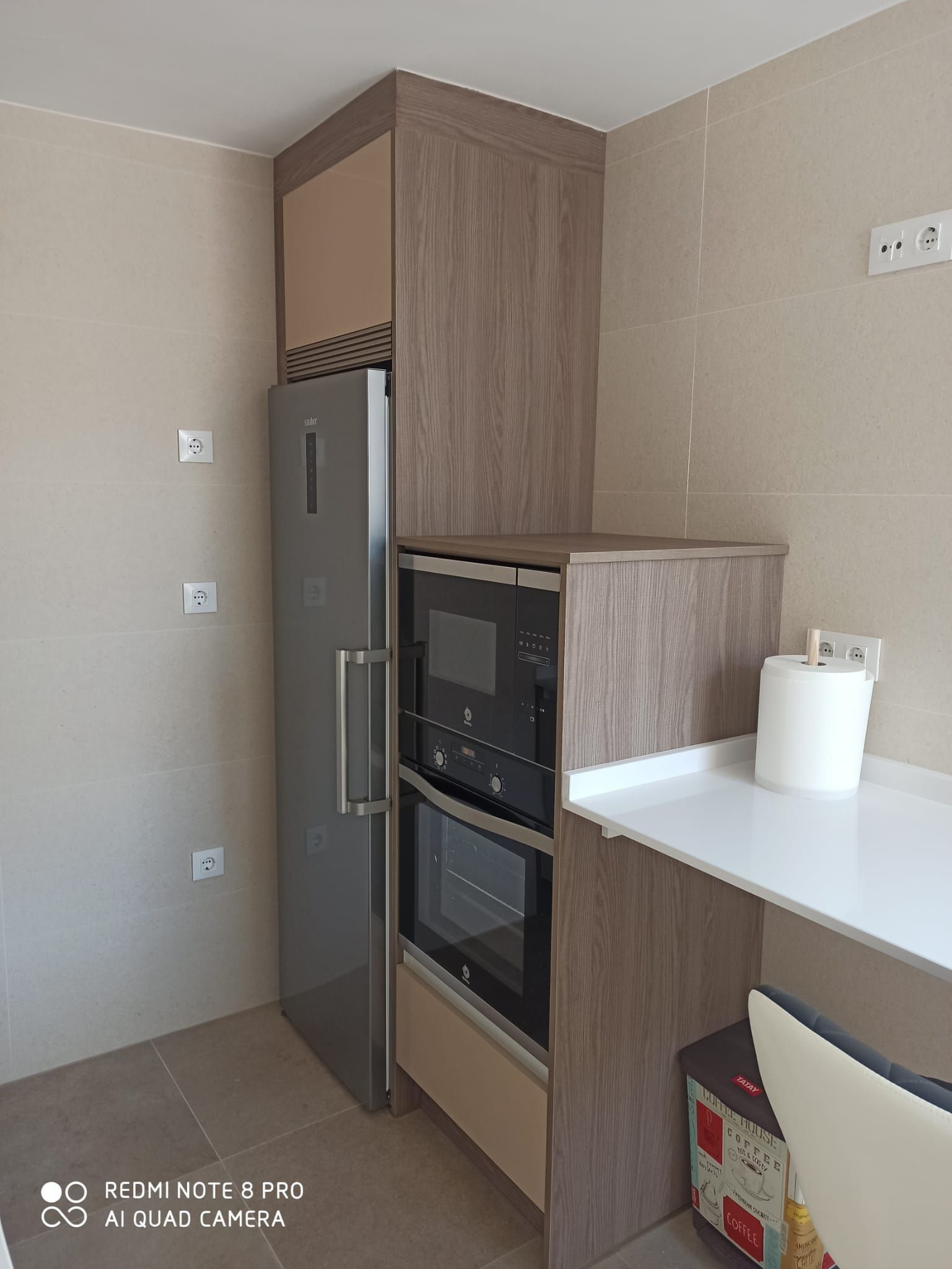 Una zona de cocina con un frigorífico alto de color gris junto a un mueble con acabado de madera que incluye un microondas empotrado y un horno de pared.