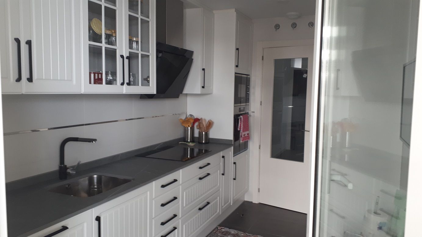 Cocina moderna blanca con encimeras gris oscuro, grifería negra y puerta con paneles de vidrio, vista desde una entrada.