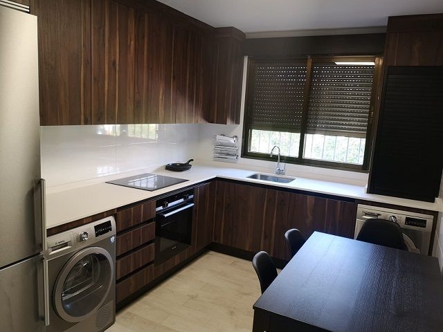 Cocina moderna con armarios de nogal, encimeras blancas, electrodomésticos de acero inoxidable y una mesa de comedor en una esquina.