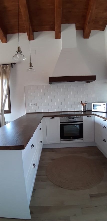 Cocina blanca en forma de U con encimera de madera oscura, salpicadero de azulejos blancos, vigas de madera en el techo y lámparas colgantes.