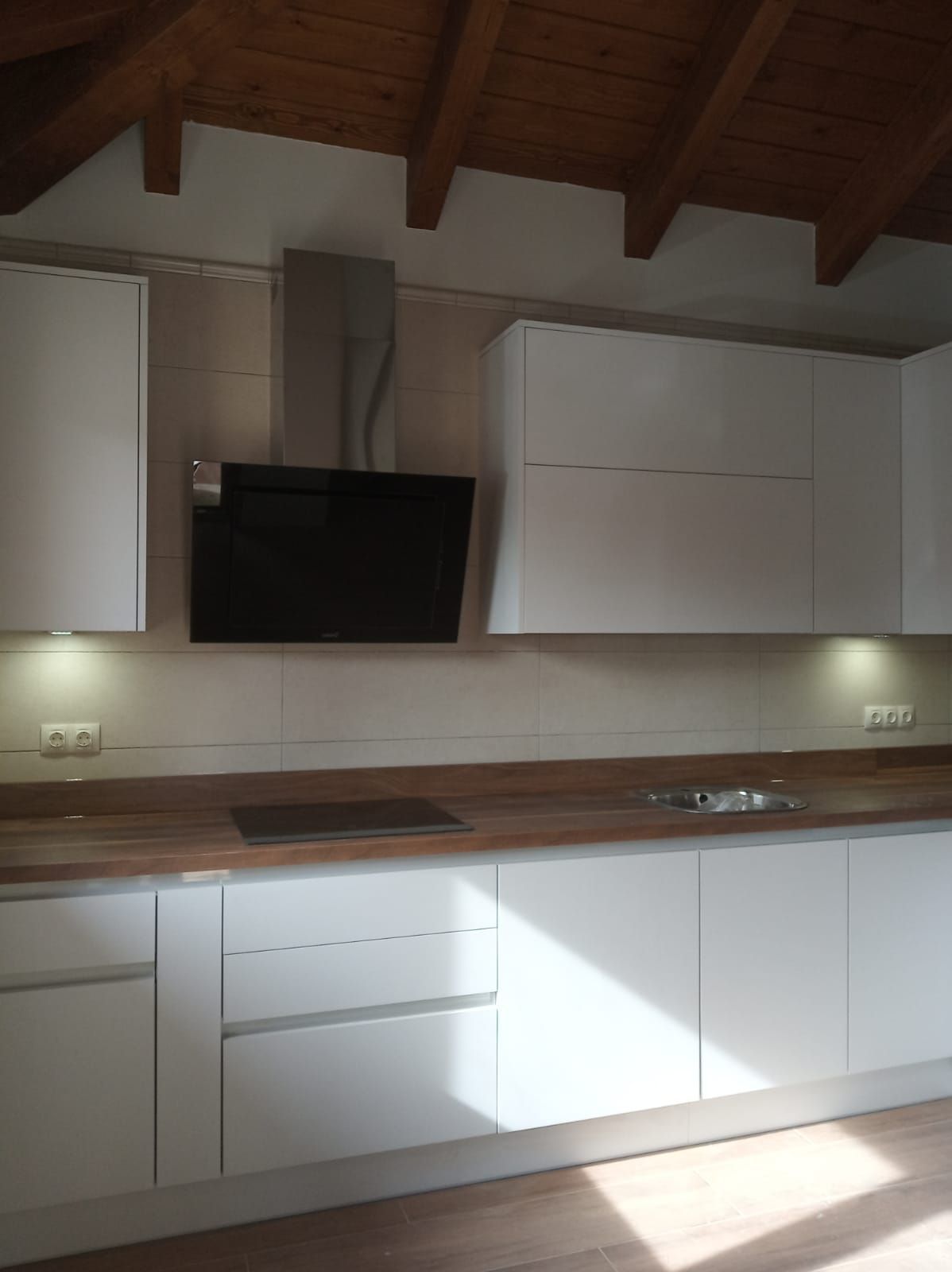 Una cocina moderna con armarios blancos, encimera de madera, campana extractora negra y vigas de madera a la vista en el techo.