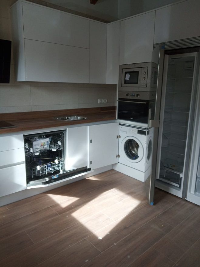 Cocina moderna con armarios blancos, suelo con aspecto de madera, lavavajillas integrado, horno y lavadora.