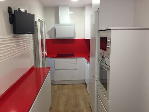 Una cocina moderna y estrecha con armarios blancos, encimeras y salpicadero de color rojo brillante, y un televisor montado en la pared.