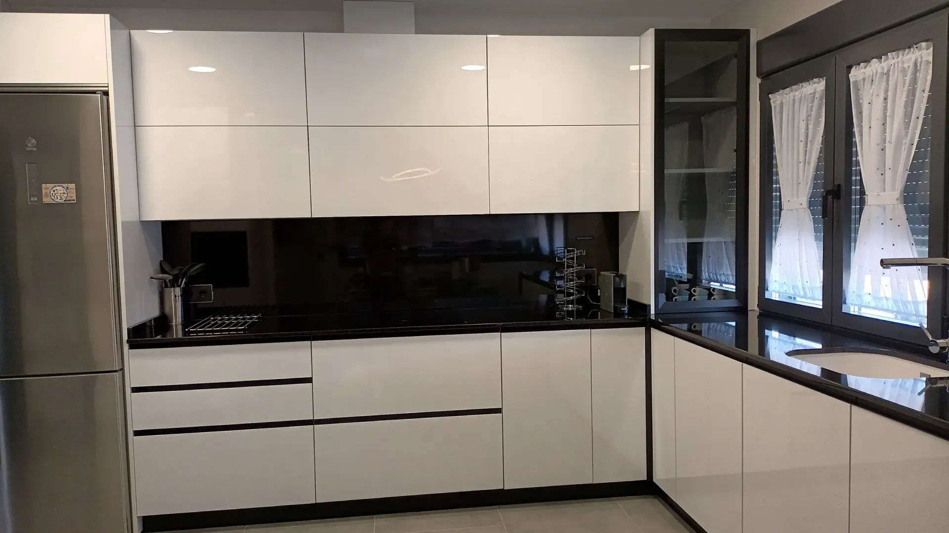 Cocina moderna con armarios blancos de alto brillo, encimera negra, frigorífico de acero inoxidable y ventana con cortinas.