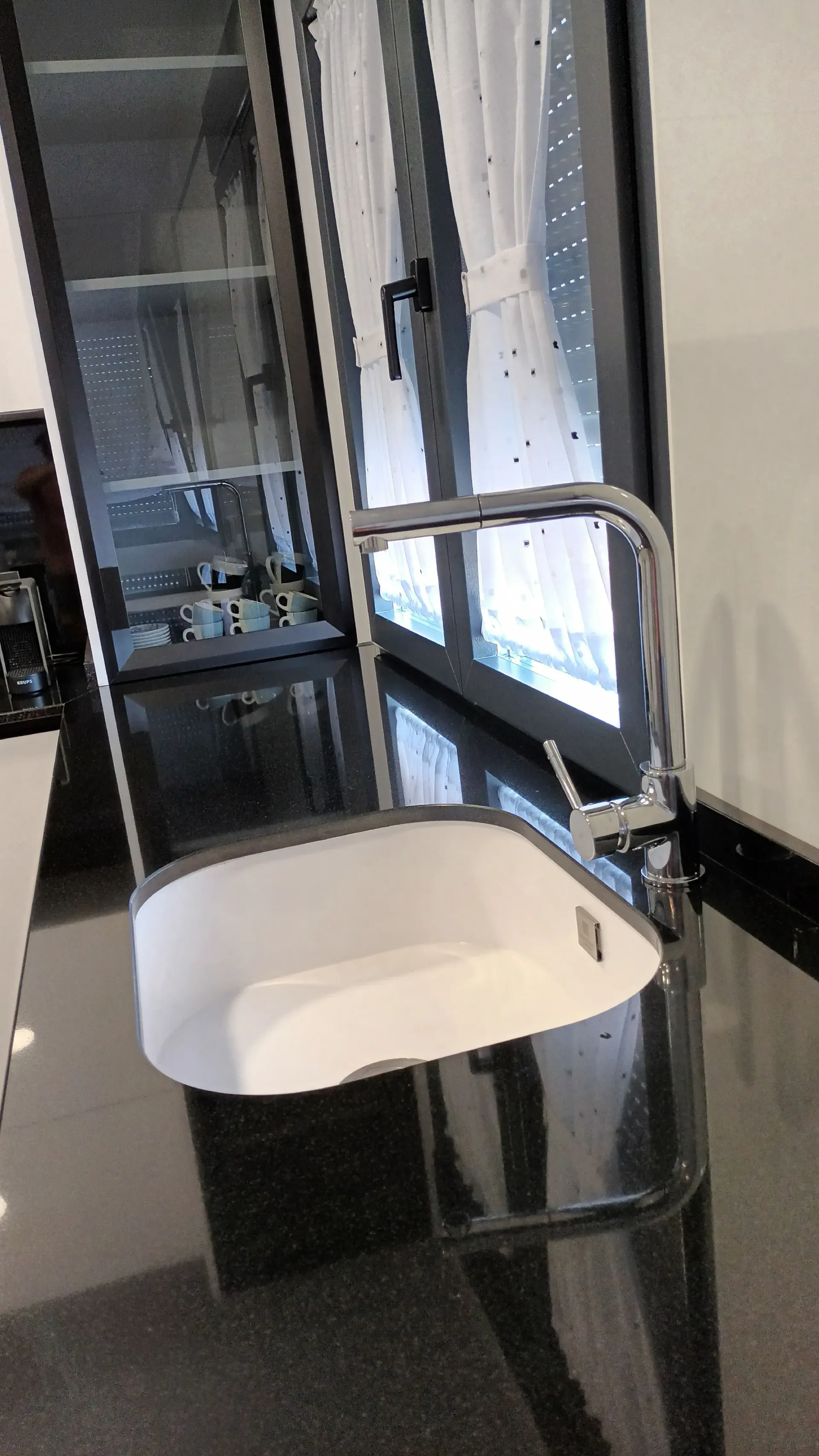 Un moderno fregadero de cocina blanco con grifo cromado, empotrado en una encimera oscura y pulida frente a una ventana.