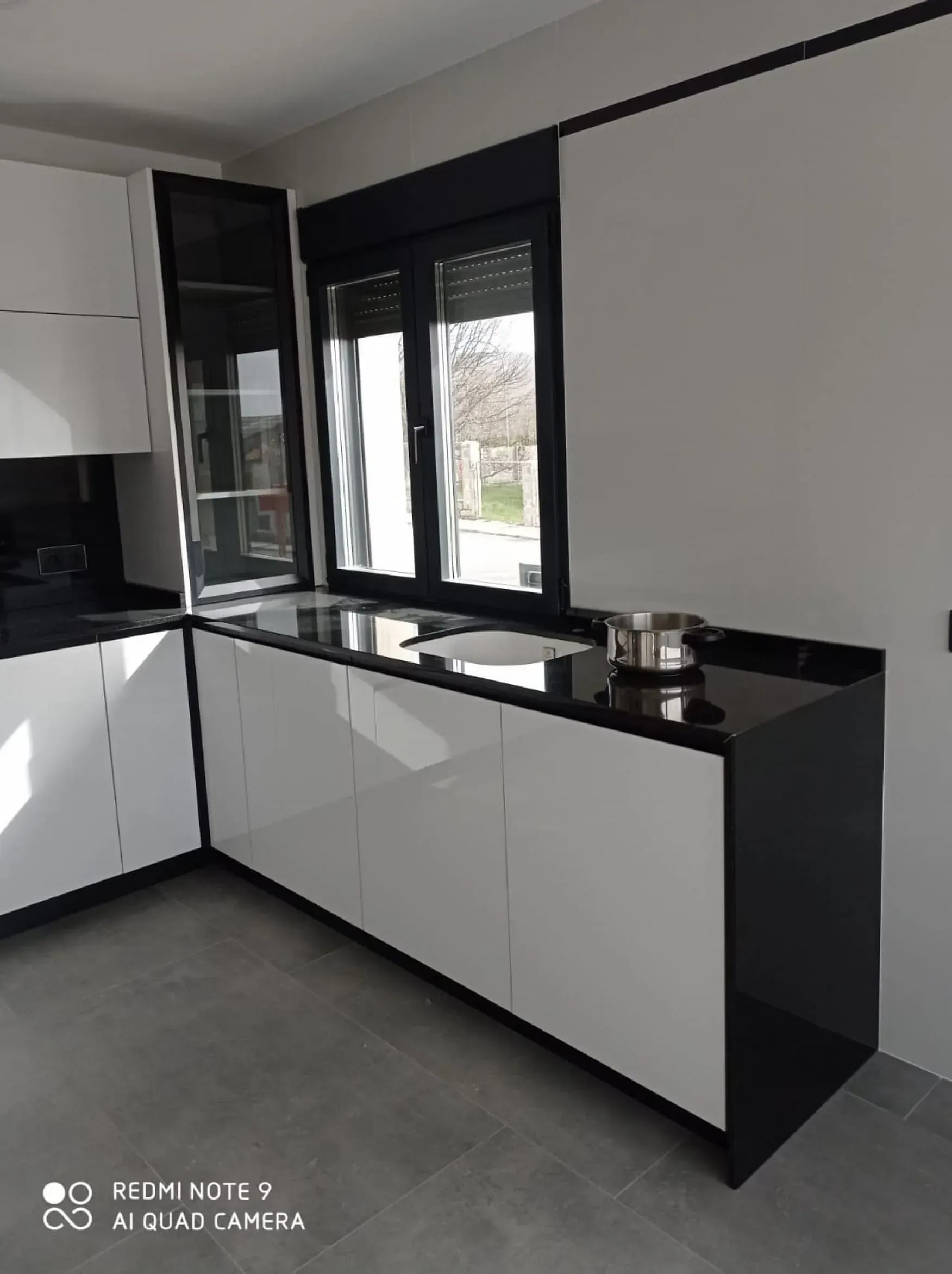 Una cocina moderna con armarios blancos brillantes, encimeras negras y un marco de ventana negro, situada sobre un suelo de baldosas grises.