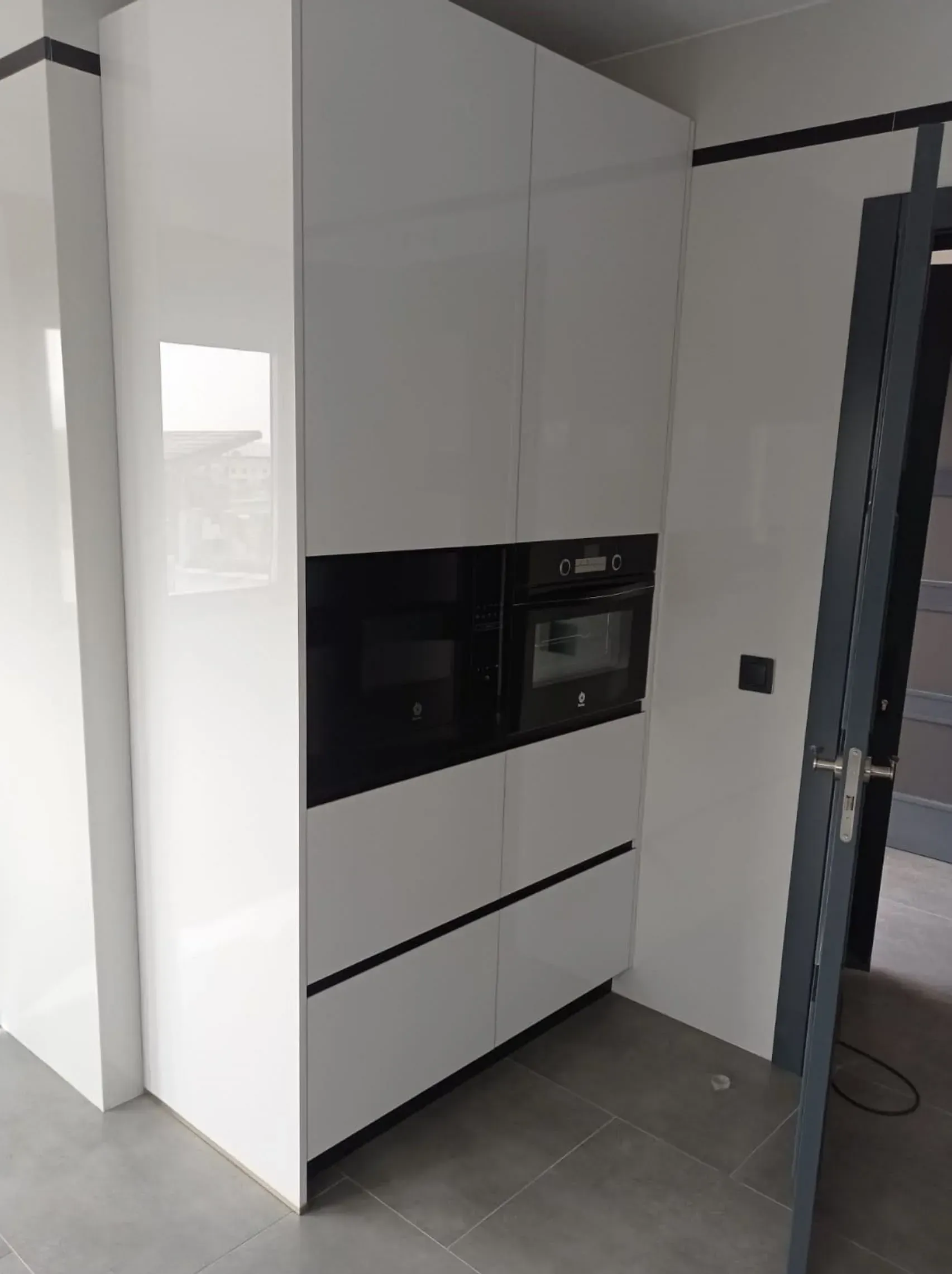 Un mueble de cocina alto, blanco y moderno, con dos hornos negros integrados, sobre un suelo de baldosas gris claro.