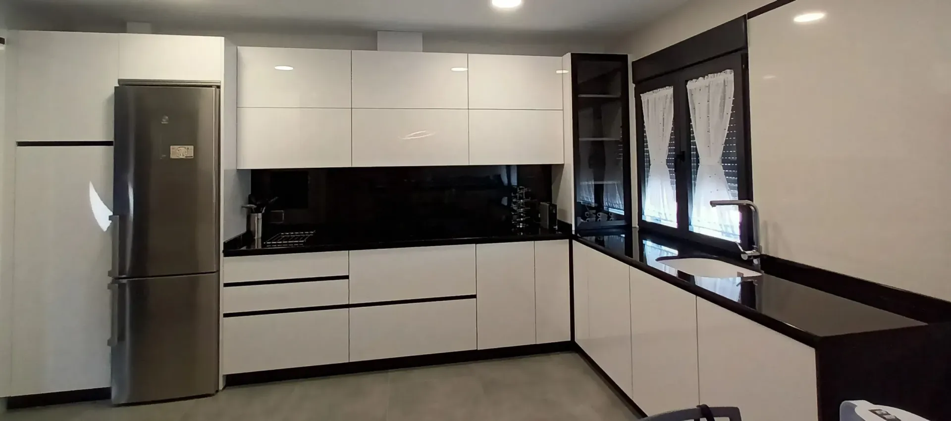 Cocina moderna en forma de L con armarios blancos brillantes, encimeras negras y electrodomésticos de acero inoxidable.