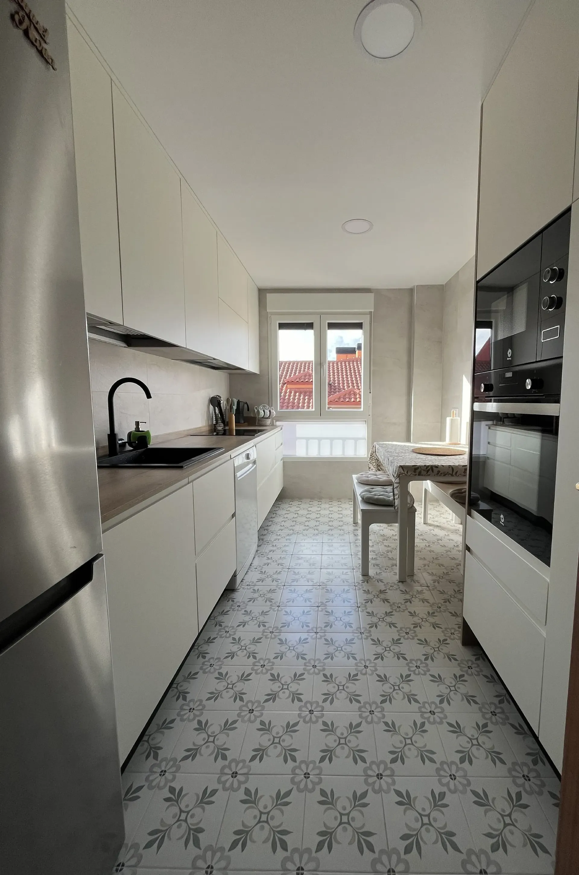 Una cocina tipo galera con gabinetes blancos, un refrigerador de acero inoxidable, electrodomésticos negros y baldosas estampadas en el piso.