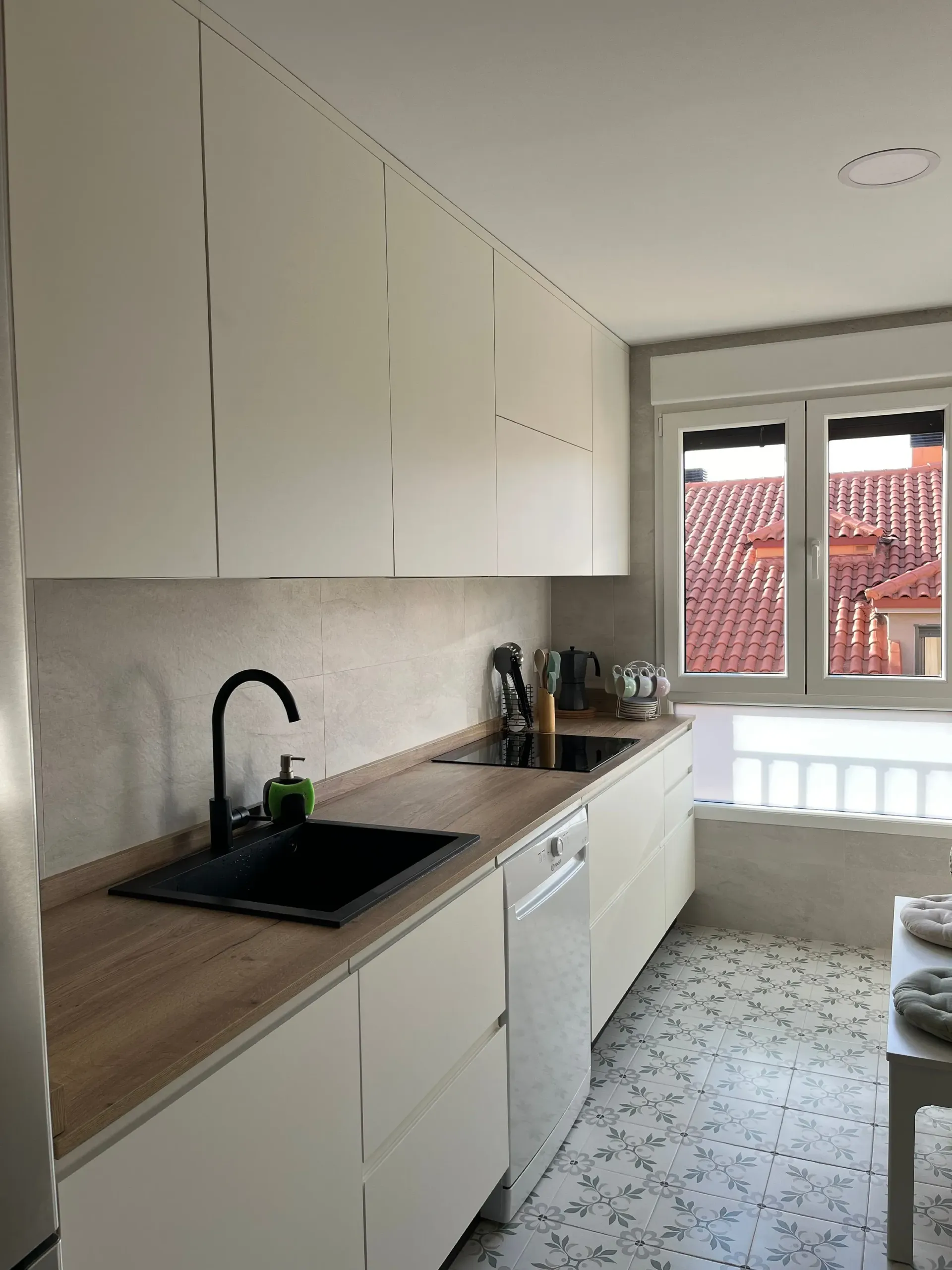 Cocina moderna tipo galera con gabinetes blancos, encimeras con apariencia de madera, fregadero negro y pisos de baldosas estampadas junto a una ventana.